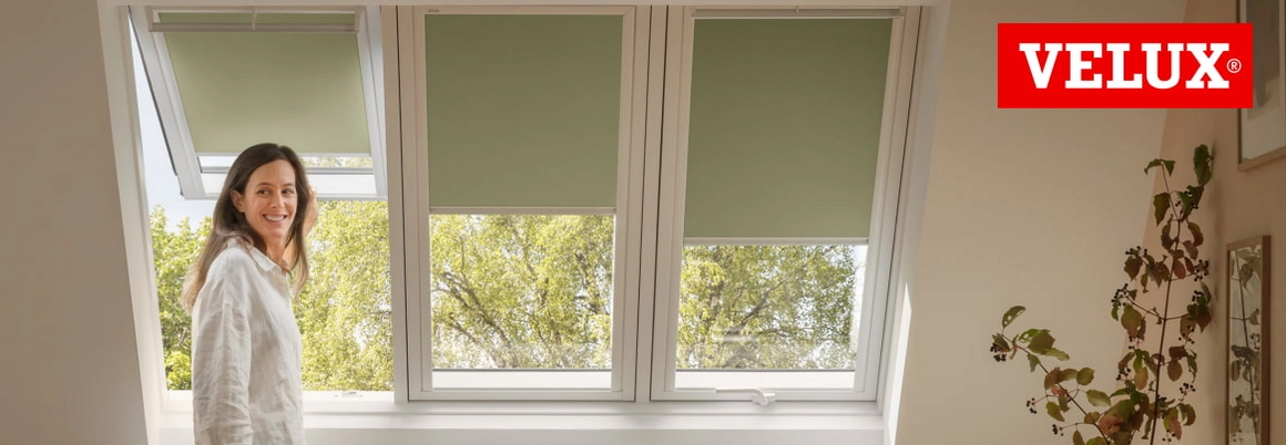 Eine Person steht lächelnd vor Dachfenstern der Marke Velux mit grünen Rollos.